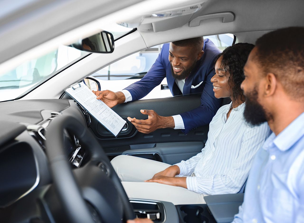 Young Black Couple Sitting Inside New Car In Showroom, Listening Professional Salesman by Prostock-studio